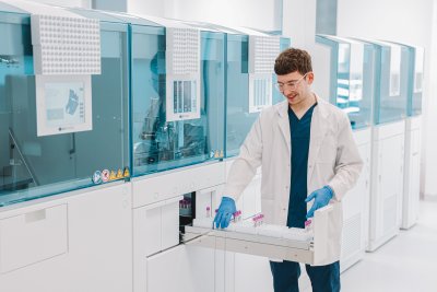 InMedica laboratorijos darbuotojas su baltu chalatu ir mėlynomis pirštinėmis deda mėgintuvėlius į automatizuotą analizatorių