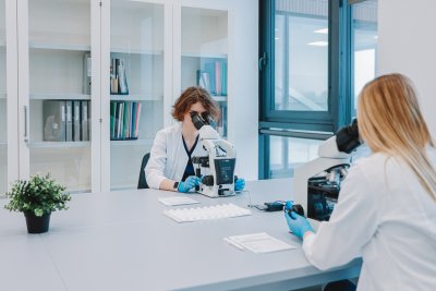 Dvi InMedica laboratorijos darbuotojos su baltomis uniformomis ir pirštinėmis tiria mėginius per mikroskopus