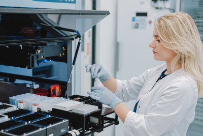 InMedica laboratorijos darbuotoja su baltu chalatu ir pirštinėmis dirba su automatizuota įranga