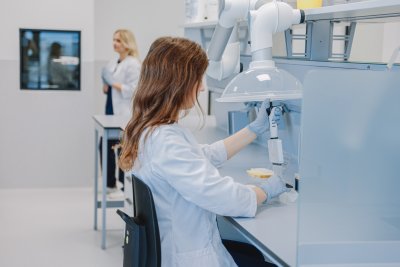 InMedica laboratorijos darbuotoja su baltu chalatu ir pirštinėmis ruošia mėginius po lokalia traukos sistema