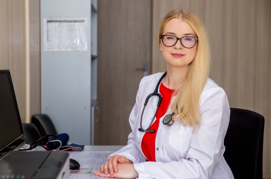 InMedica gydytoja su baltu chalatu ir stetoskopu sėdi prie darbo stalo kabinete