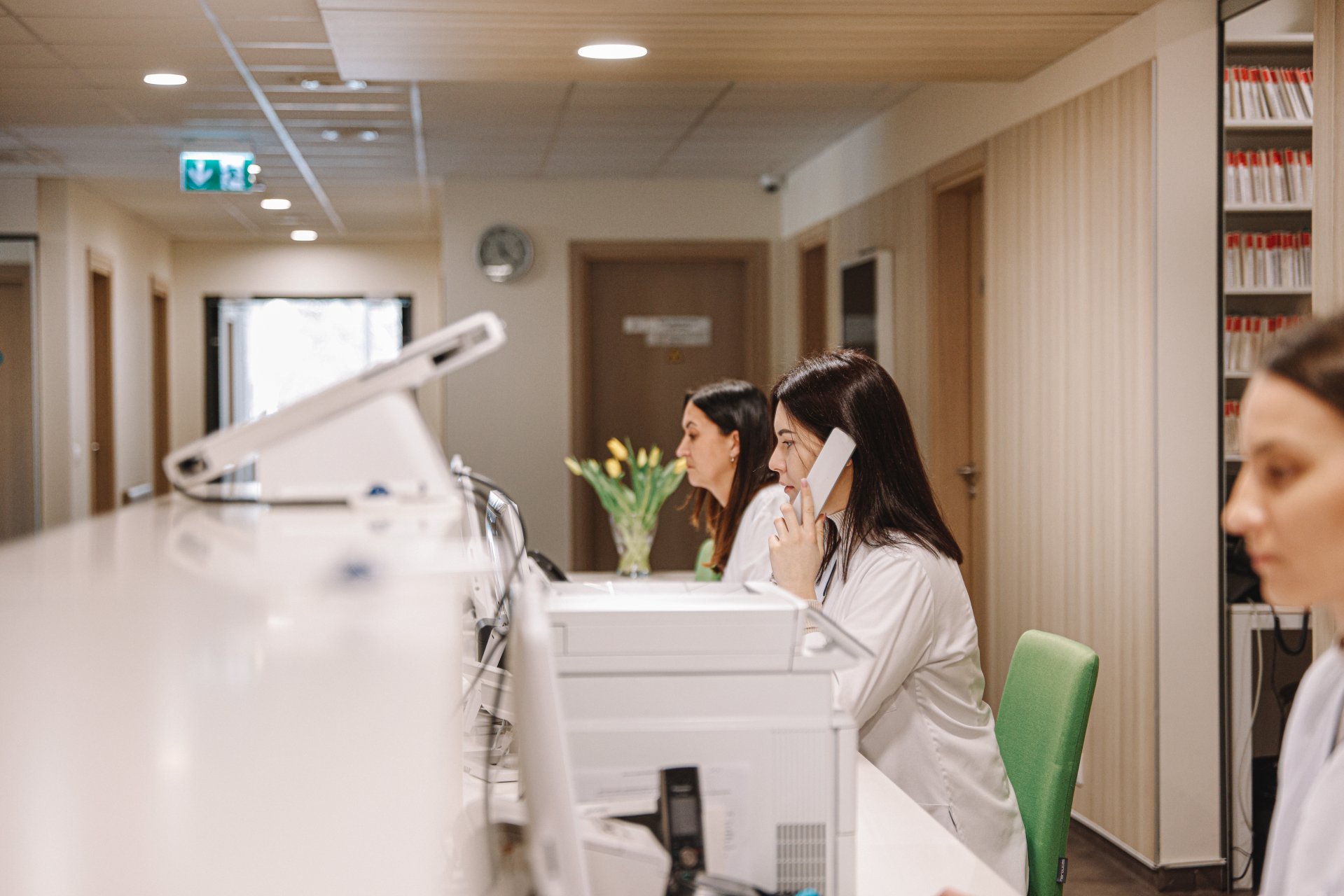 InMedica registratūros darbuotojos dirba kompiuteriu ir telefonu moderniame klinikos priimamajame